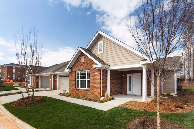 Single-story brick and siding cottage-style building with a covered porch, attached garage and landscaped lawn under a blue sky.
