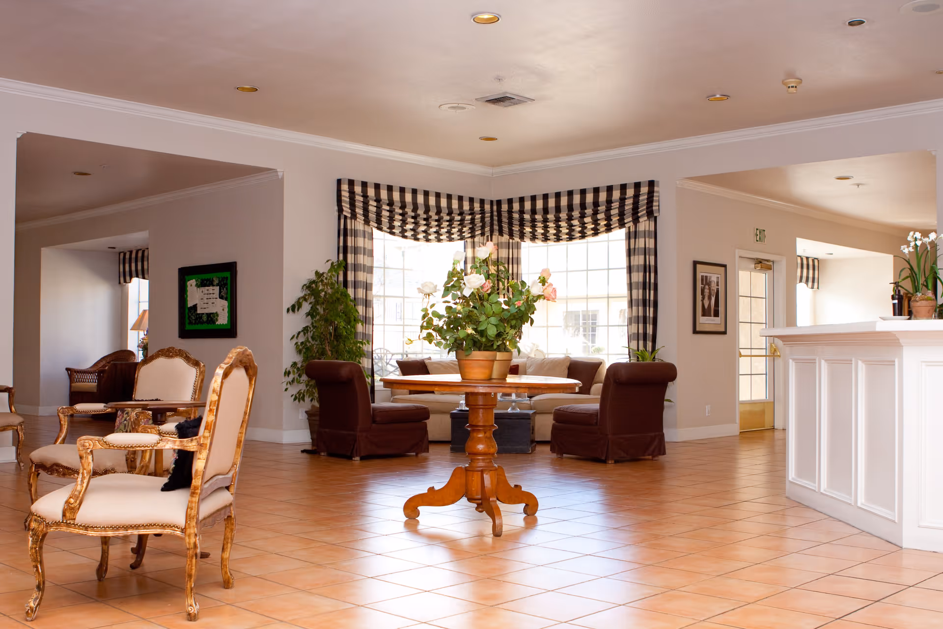 A spacious and well-lit assisted living community common area with tiled floors, a round wooden table with a potted plant in the center, several upholstered chairs, large windows with checkered valances, and a white reception desk on the right side.
