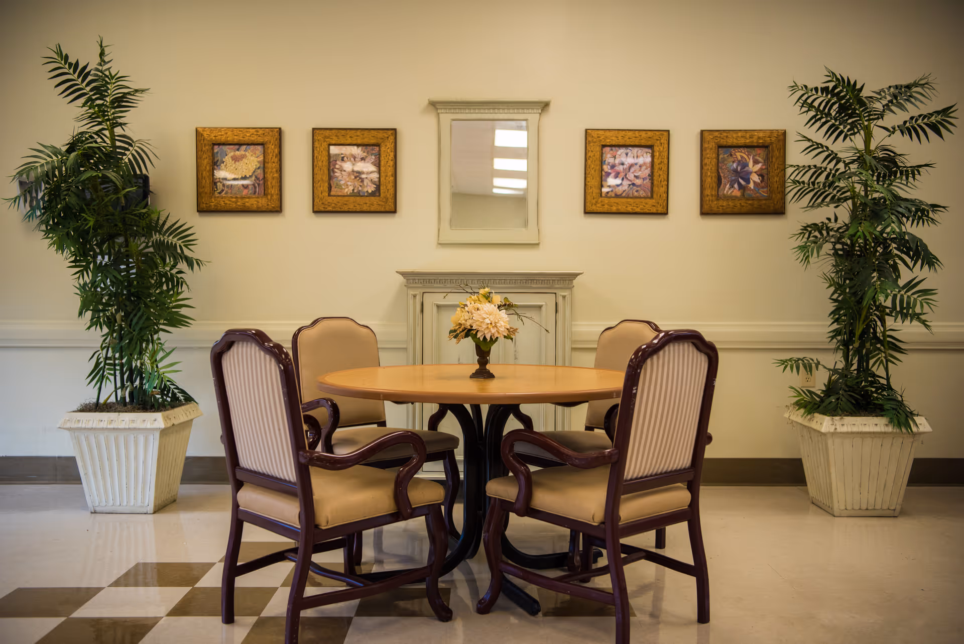 A round wooden table with a floral centerpiece surrounded by four upholstered chairs with wooden frames in a room. The wall behind features four framed floral paintings and a decorative mirror above a small white mantel. Two large potted plants flank the table on either side.