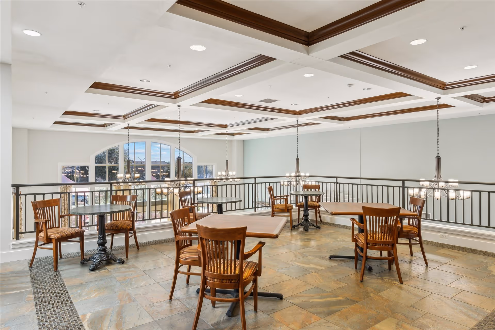 A bright and spacious dining area with several wooden tables and chairs arranged on a tiled floor. The ceiling features decorative wooden beams and hanging light fixtures. Large windows allow natural light to fill the space, and a black metal railing borders the area.