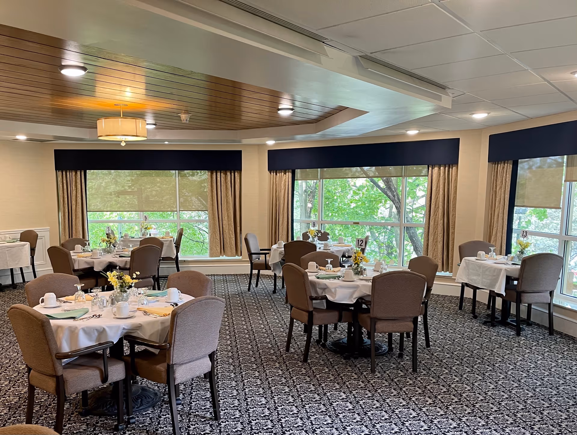 A dining room with several round tables covered with white tablecloths, each set with cups, glasses, napkins, and small flower arrangements. The room has large windows with beige curtains and dark valances, allowing natural light to brighten the space. The ceiling features recessed lighting and a wooden accent section.