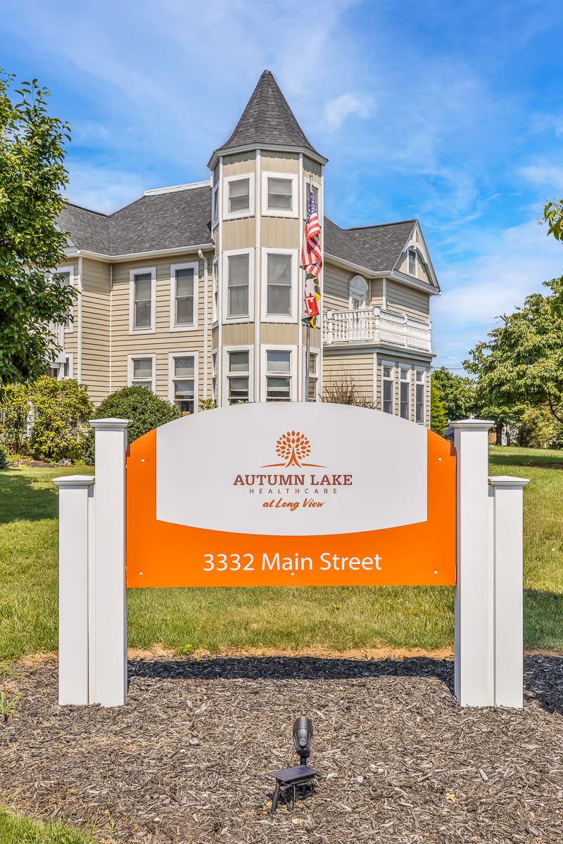 Exterior view of a beige multi-story building with a turret and white trim, behind a sign that reads 'Autumn Lake Healthcare at Long View, 3332 Main Street' on a sunny day with a blue sky and some trees around.