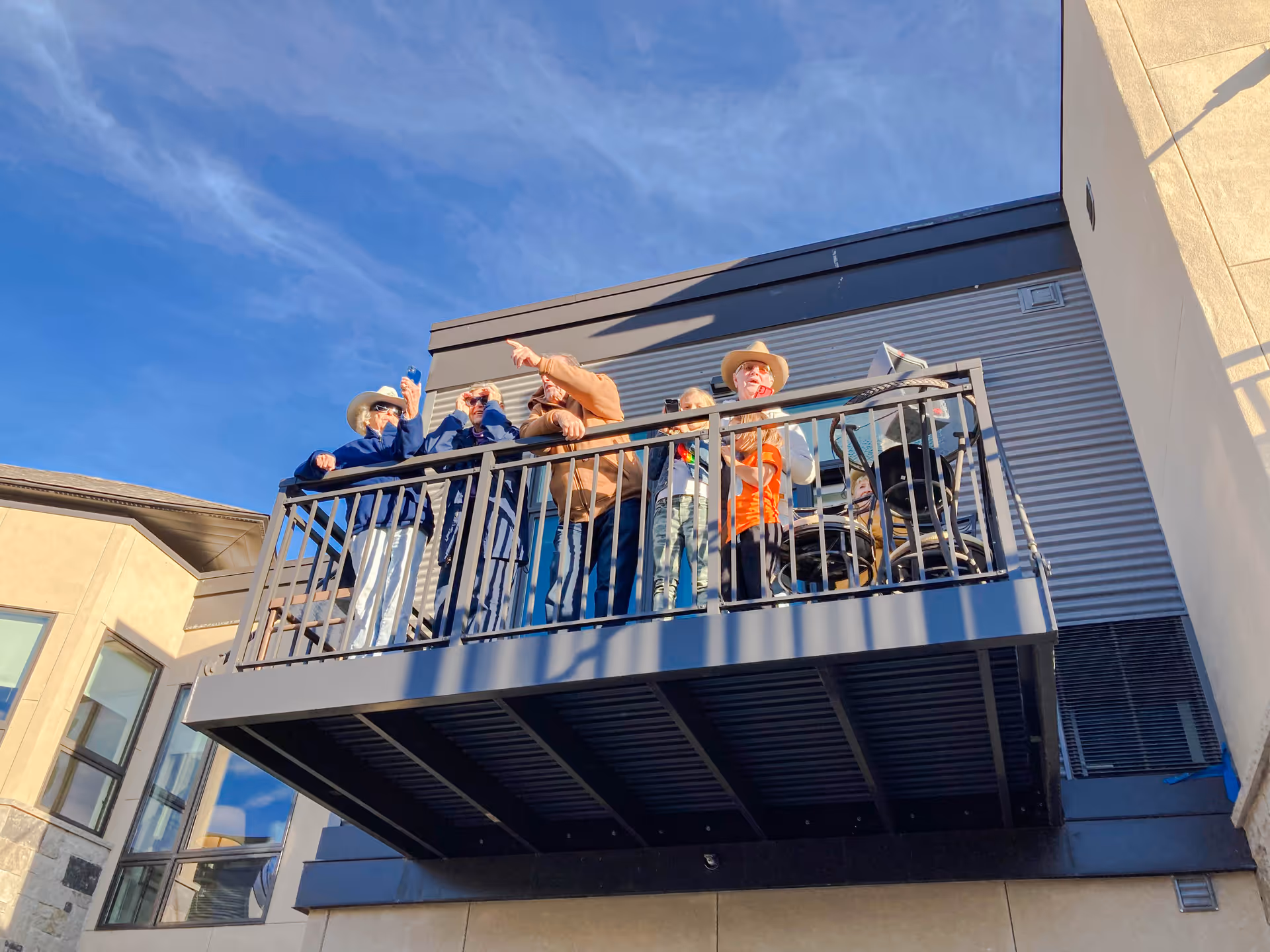 A group of elderly people standing and sitting on a balcony of a senior living facility under a clear blue sky. Some are wearing hats and sunglasses, and one person is in a wheelchair.