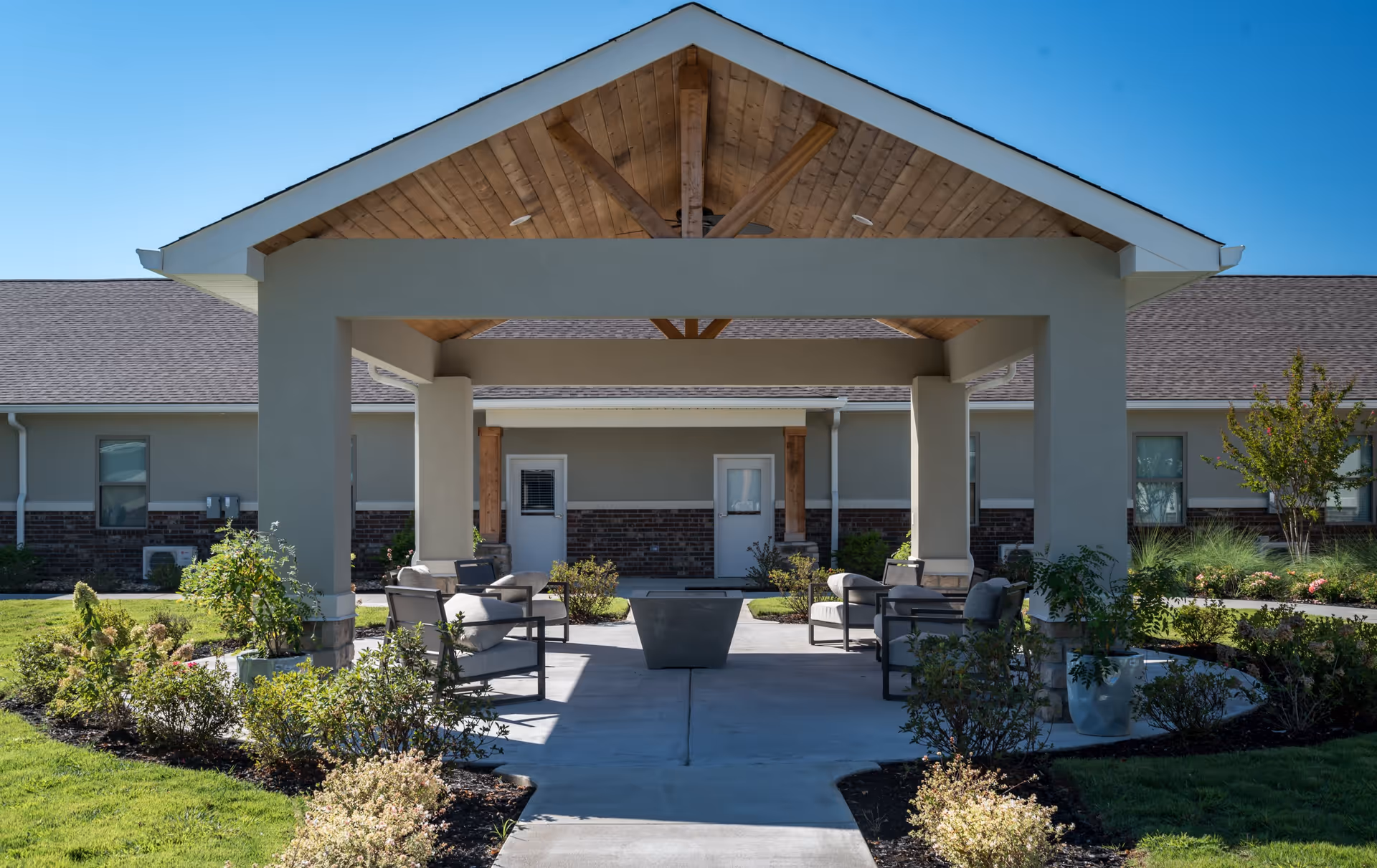 Outdoor covered seating area at Crestview Senior Living with cushioned chairs arranged around a central fire pit, surrounded by landscaped greenery and a building with doors and windows in the background under a clear blue sky.