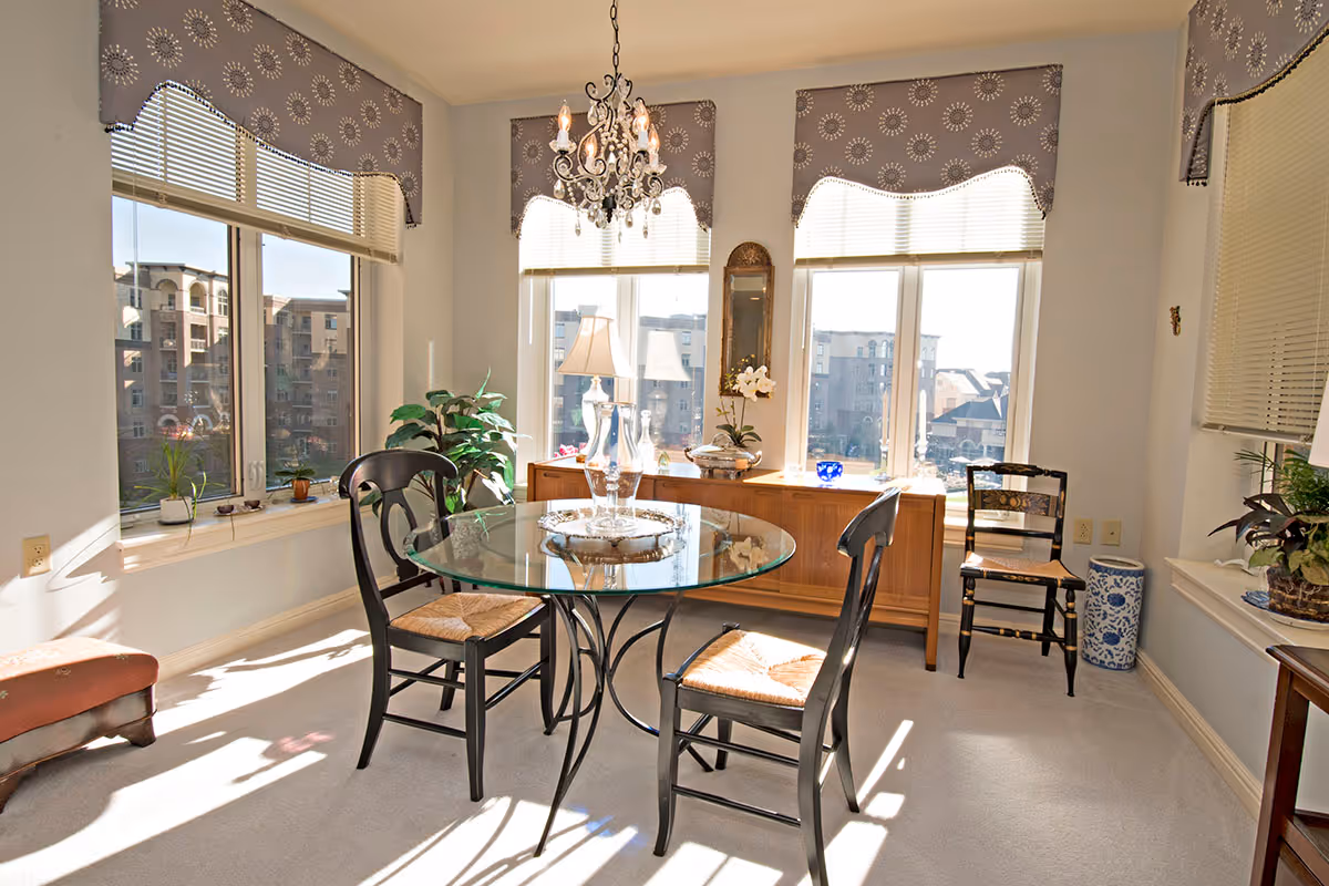 A bright dining area with a round glass table and four black wooden chairs with woven seats. The room has large windows with patterned valances, letting in natural light and showing a view of nearby buildings. There is a chandelier hanging above the table, a wooden sideboard with decorative items, and several potted plants around the room.