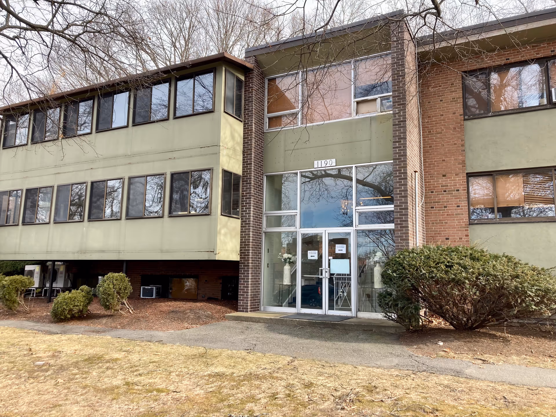 Front entrance of a three-story mid-century brick and green-paneled building with glass double doors numbered 1190 and shrubs in front.