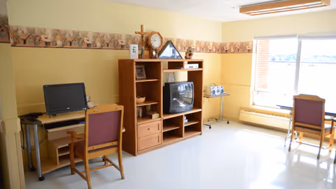 Bright communal living room with a TV cabinet, desks and chairs near a large window.