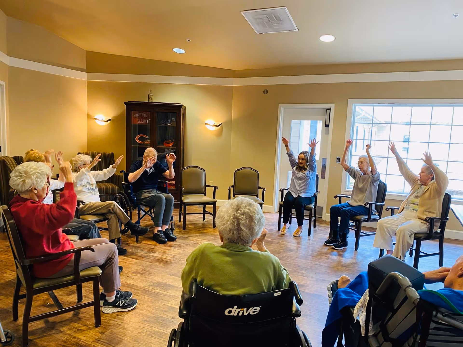 A group of elderly people and a caregiver sitting in a circle in a well-lit room with wooden floors, participating in a seated exercise or activity with their arms raised. Some individuals are in wheelchairs, and the room has beige walls, a cabinet with decorations, and large windows letting in natural light.
