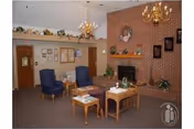 Cozy senior living common room with a brick fireplace, several armchairs, wooden tables, chandeliers, and decorative plants.