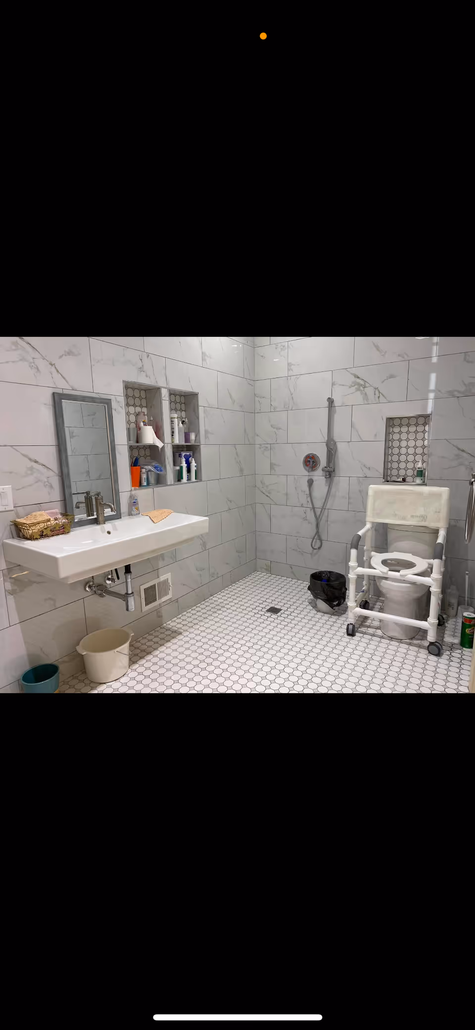A clean, accessible bathroom with white hexagonal floor tiles and large white marble wall tiles. The room features a wall-mounted sink with a mirror above it, two recessed shelves holding toiletries, a handheld showerhead, and a raised toilet with support rails for accessibility. There are also two small buckets on the floor and a black trash bin near the toilet.