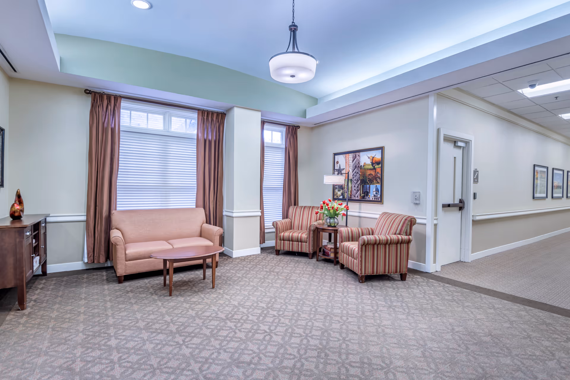 A cozy seating area in a senior living facility with a beige loveseat, two striped armchairs, a wooden coffee table, and a side table with a vase of red flowers. The room has large windows with brown curtains, patterned carpet, soft lighting from a ceiling fixture, and framed artwork on the walls. A hallway with additional framed pictures and a closed door is visible to the right.
