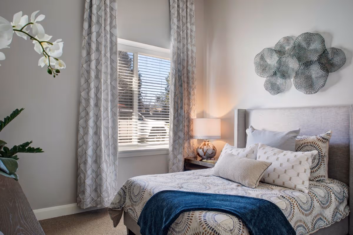 A cozy bedroom with a bed featuring patterned bedding and multiple pillows. There is a bedside table with a lamp, a window with patterned curtains, and decorative wall art above the bed. A white orchid plant is partially visible on the left side.