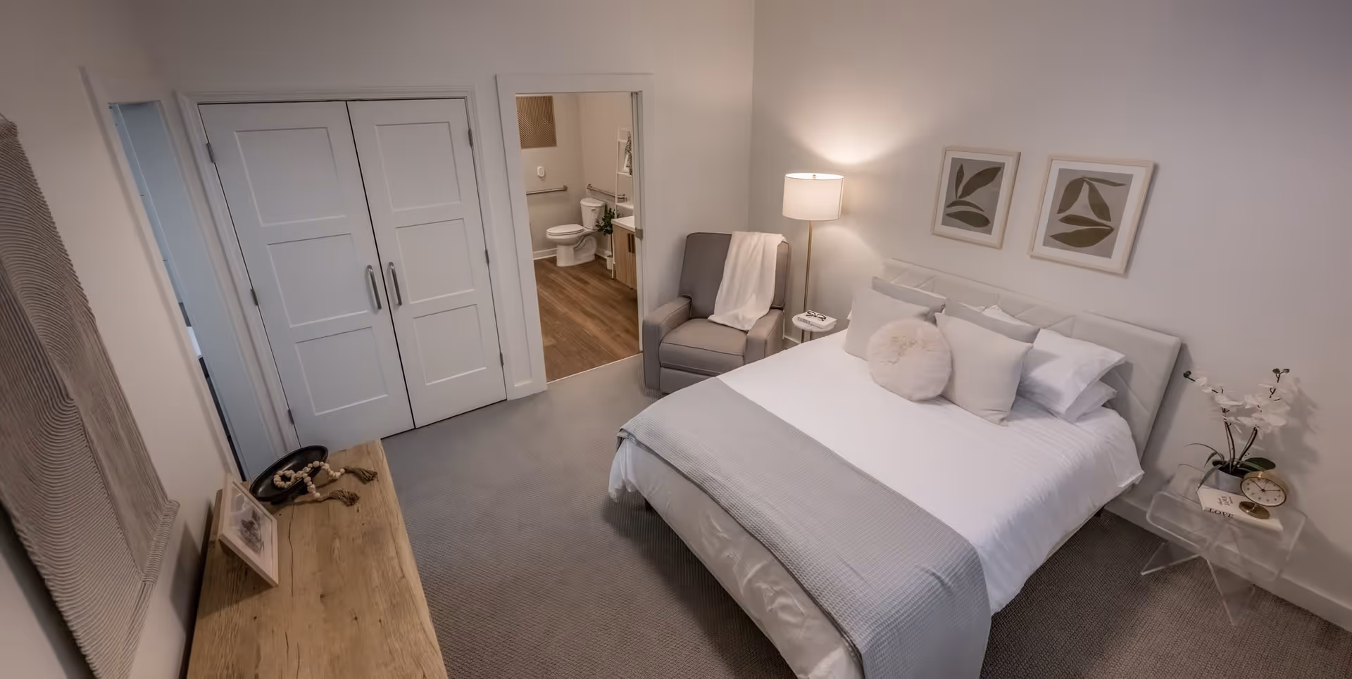 A cozy bedroom in a senior living facility featuring a neatly made bed with white and gray bedding, several pillows, and a round decorative cushion. There is a gray armchair with a white throw blanket next to a floor lamp and a small side table with a book. On the right side, a clear acrylic side table holds a clock, a book, and a white orchid plant. The room has light-colored walls with two framed leaf prints above the bed. A wooden dresser with decorative items is on the left side, and a doorway leads to an adjacent bathroom with a toilet and vanity visible.