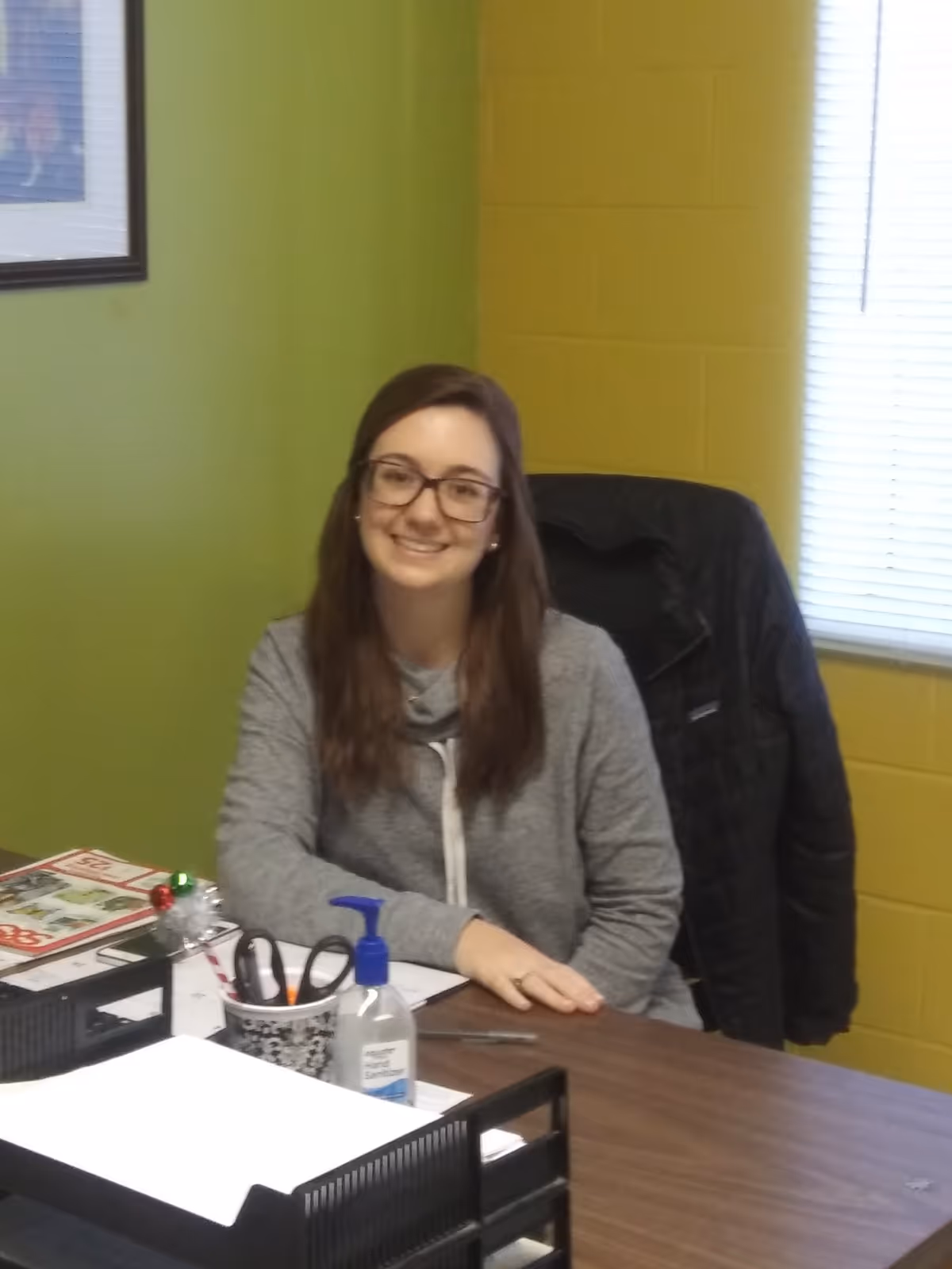 A woman with long brown hair and glasses is sitting at a desk in an office with green and yellow walls. On the desk are office supplies including a pen holder, hand sanitizer, papers, and a magazine. A black jacket is draped over the back of her chair, and a window with blinds is visible behind her.