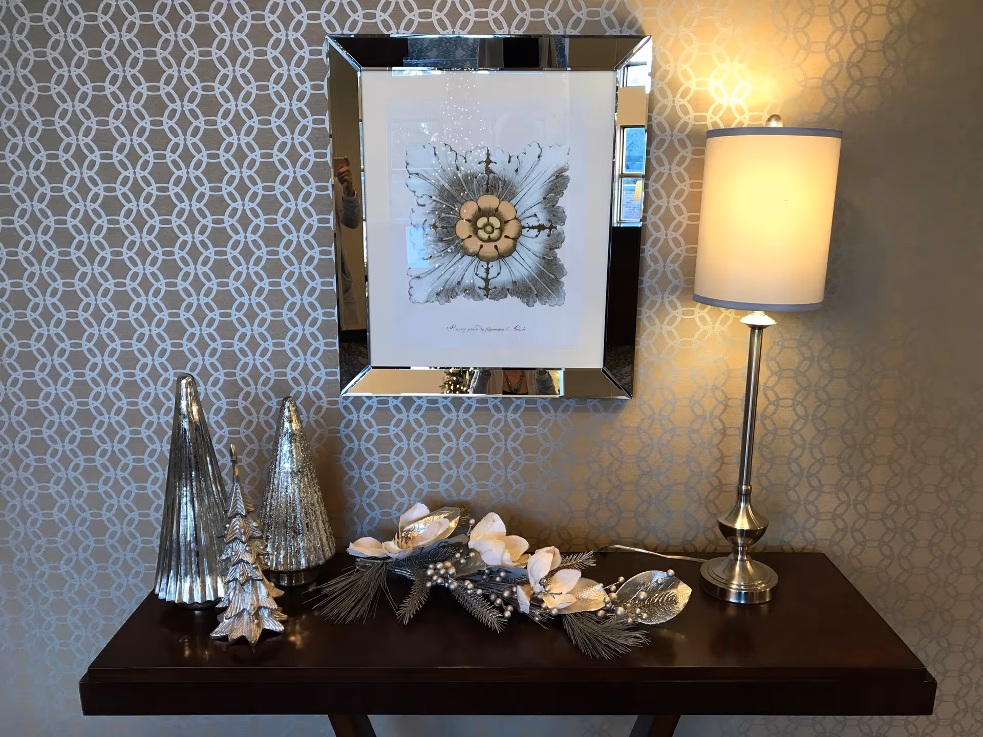A decorative console table against a patterned wall with a framed floral artwork above it. On the table are three metallic cone-shaped sculptures resembling trees, a floral and pine cone arrangement, and a silver table lamp with a white lampshade.