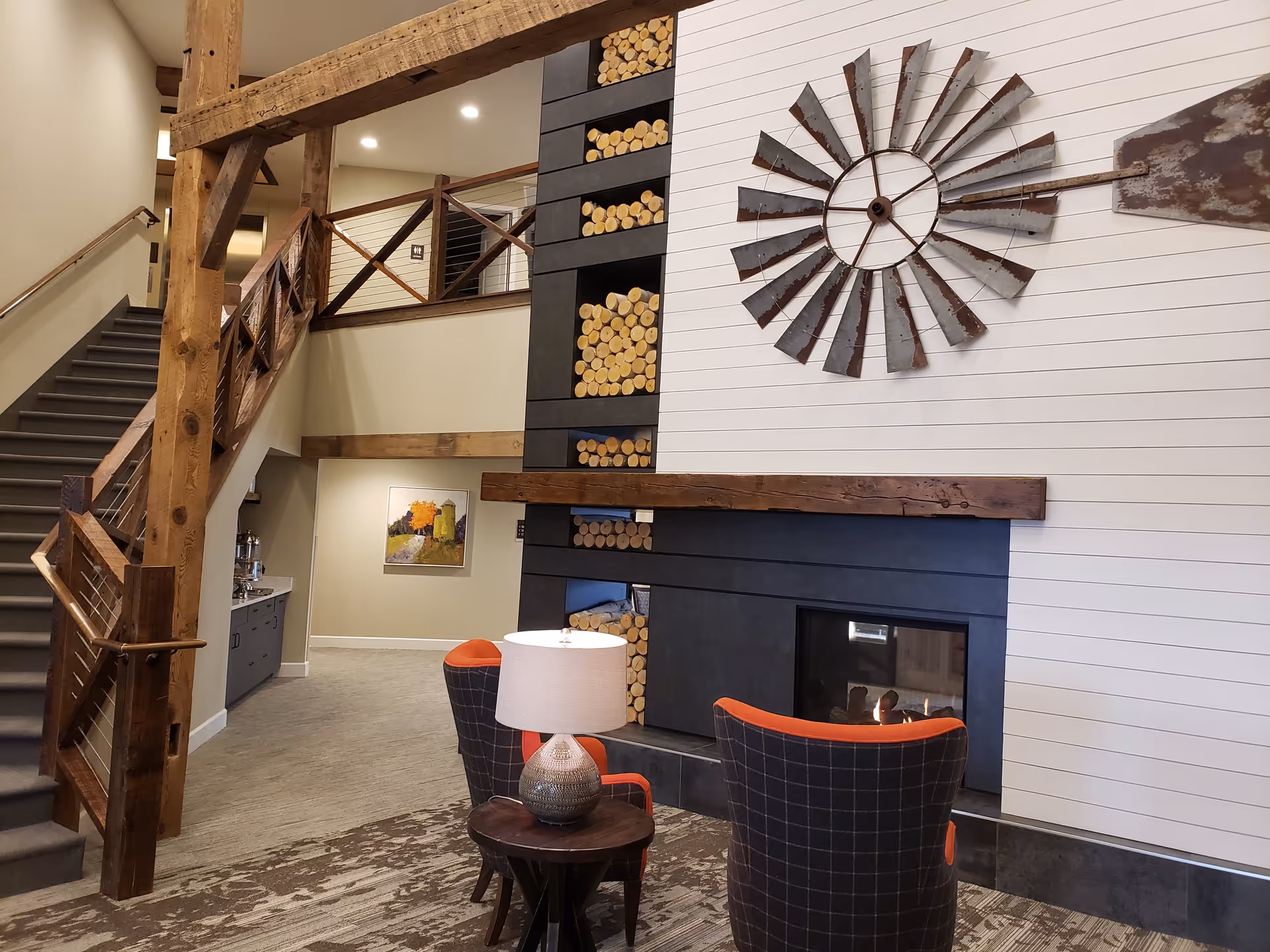 Interior view of a senior living facility common area featuring a modern fireplace with stacked firewood, two plaid armchairs with orange accents, a small round wooden table with a lamp, wooden beams, a staircase with wooden railing, and a painting on the wall in the background.
