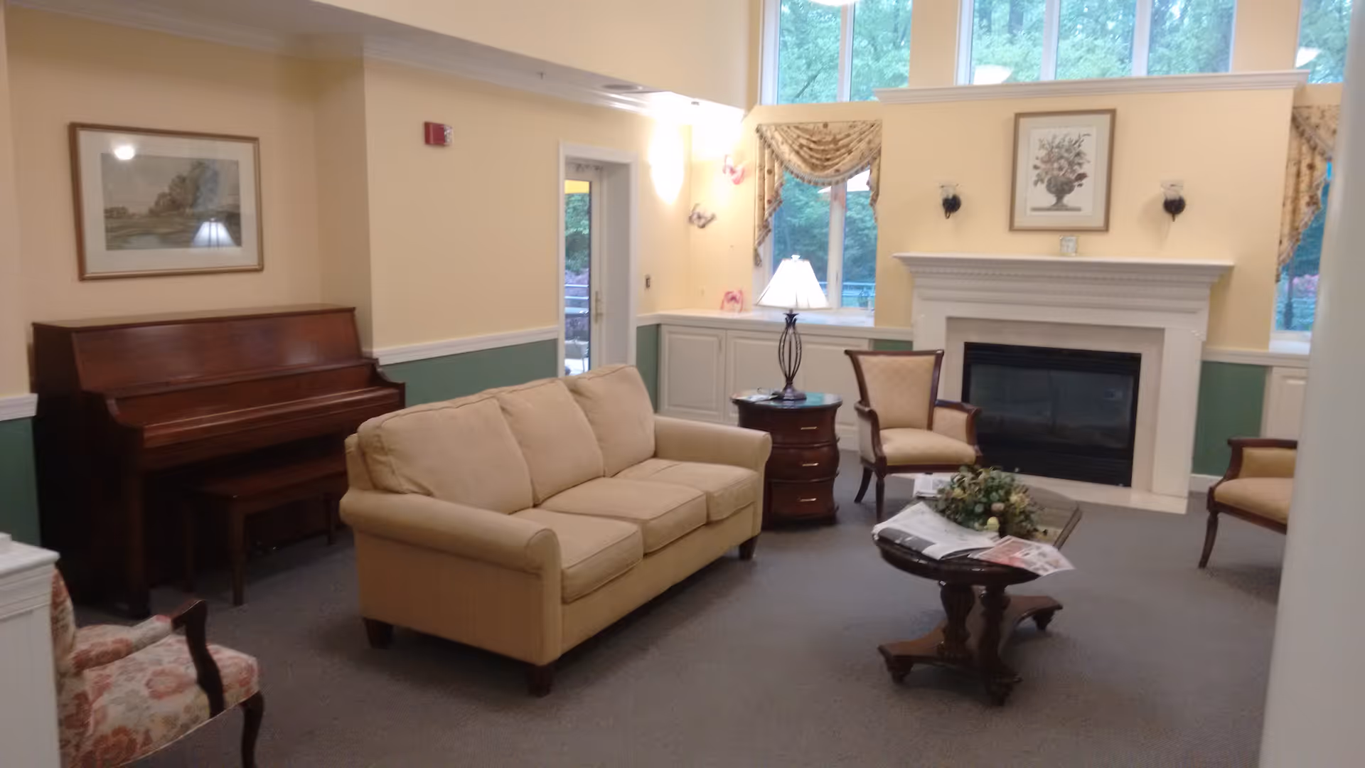 A cozy living room with a beige sofa, two upholstered chairs, a wooden coffee table with a floral centerpiece and newspapers, a side table with a lamp, a fireplace with a framed floral picture above it, a wooden piano with a bench, and windows with decorative curtains letting in natural light.