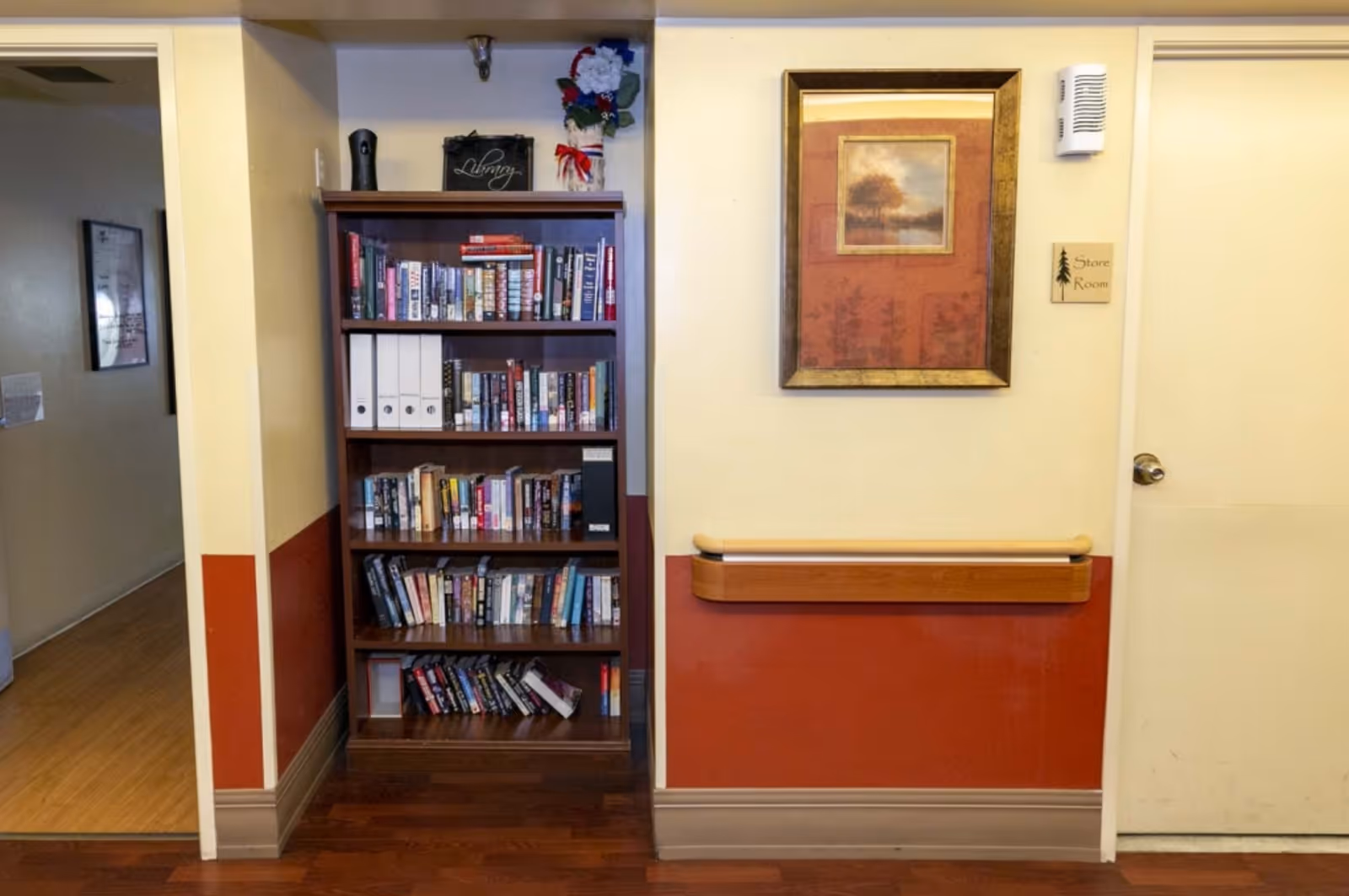 A hallway corner in Holladay Healthcare Center featuring a wooden bookshelf filled with books and binders. To the right, there is a door labeled 'Store Room' with a framed picture of a tree hanging above a wooden handrail. The walls are painted beige and reddish-brown, and the floor is wooden.