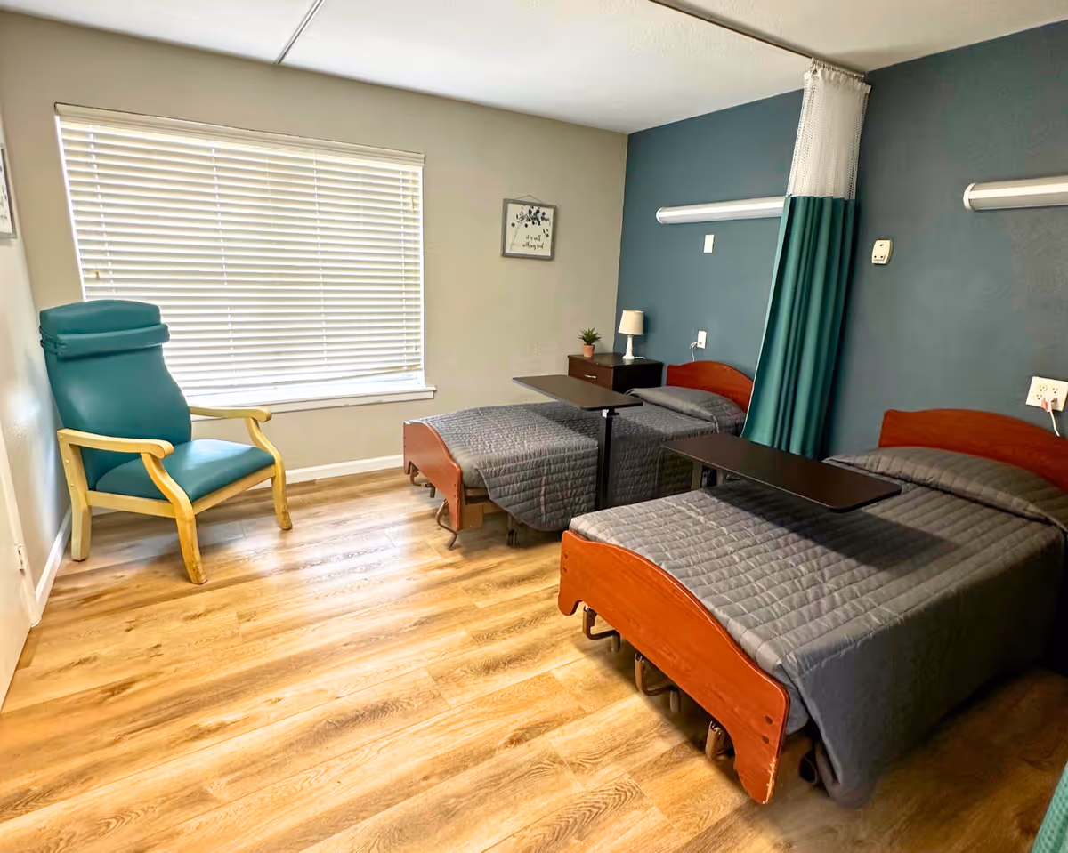 A room with two single beds covered with gray quilts, each with an overbed table. There is a green and wood armchair near a window with closed blinds. A small nightstand with a lamp and a small plant is placed between the beds. The walls are painted in beige and dark teal, and a privacy curtain hangs between the beds. The floor is wood-patterned.