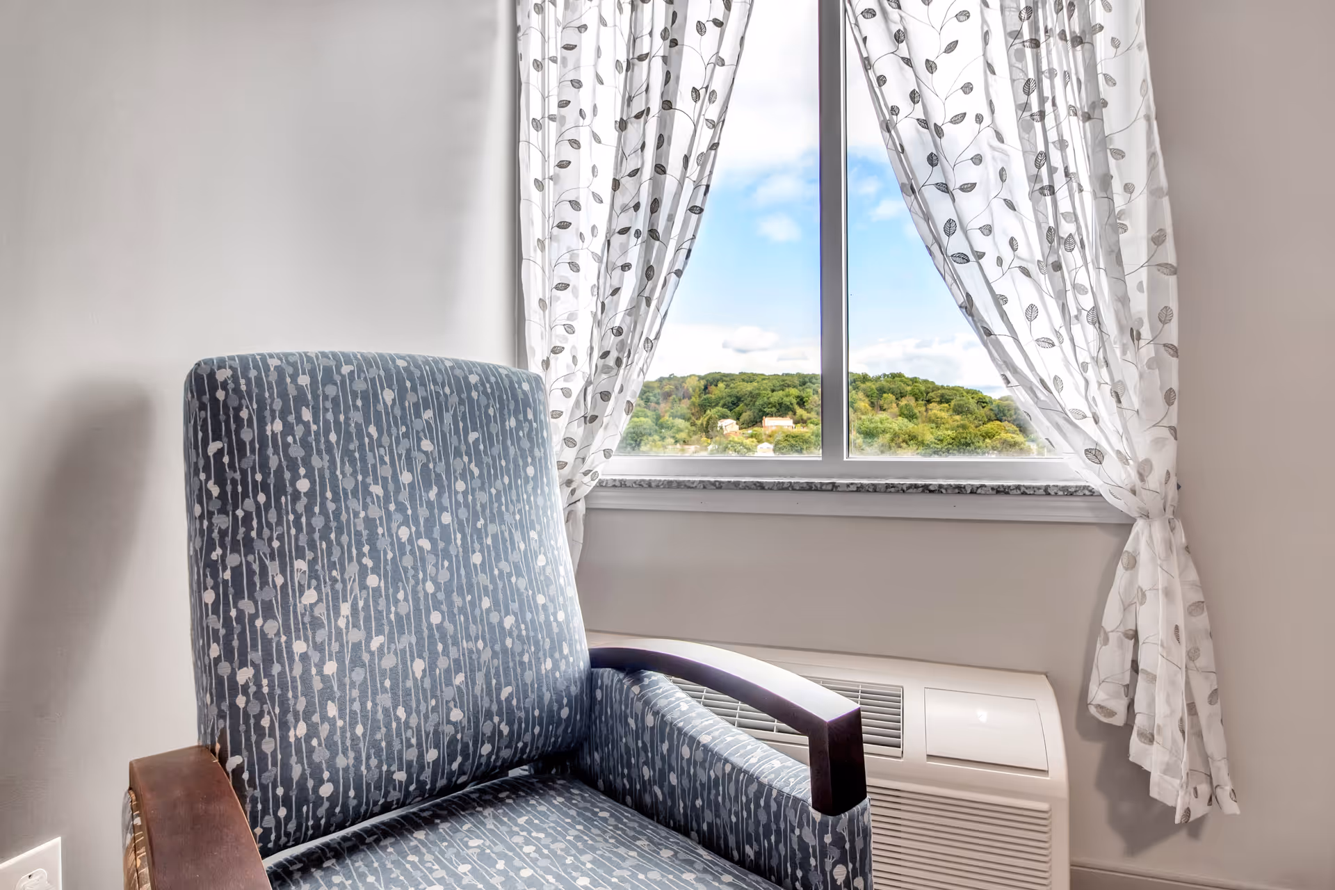 Patterned upholstered armchair beside a curtained window with a scenic view and an air conditioning unit below.