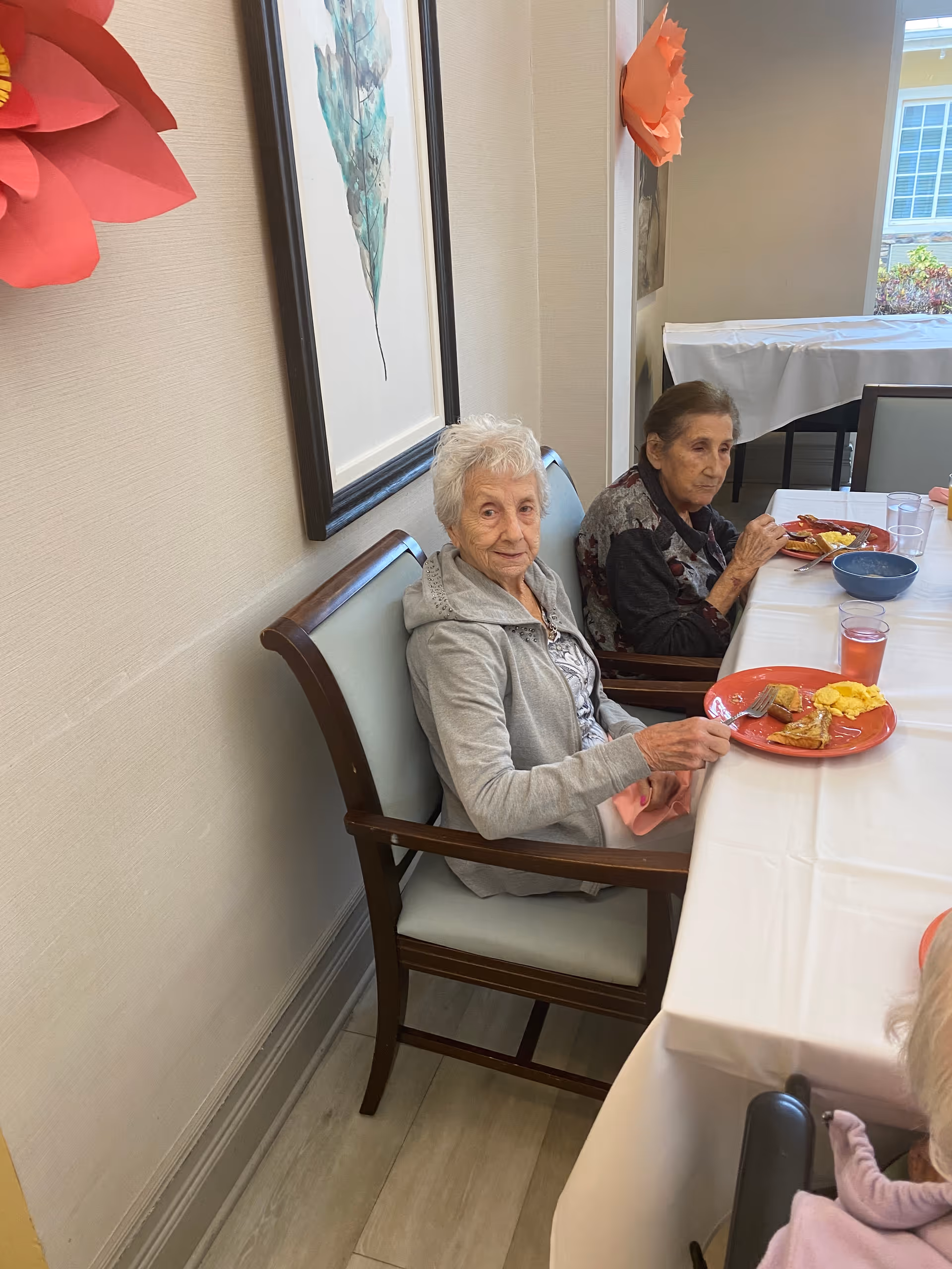 Two elderly women sit at a dining table in a senior living facility eating from red plates.