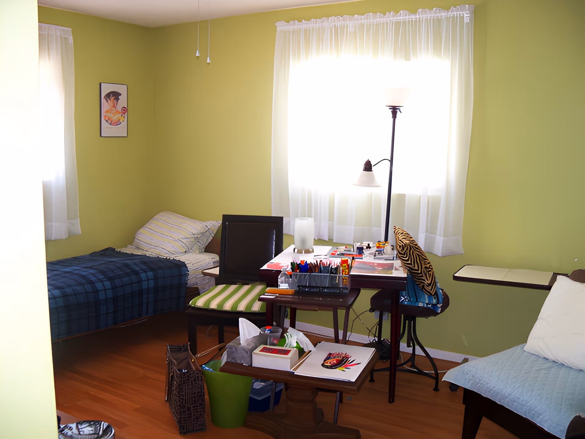 A small bedroom with light green walls and wooden floor. There is a single bed with striped bedding and a blue plaid blanket on the left side. A window with sheer white curtains lets in bright light. In front of the window is a desk cluttered with art supplies, papers, and a lamp. Two chairs are positioned near the desk, one with a green striped cushion and the other with a zebra-patterned pillow. A small table with tissues and art materials is also visible. A framed picture hangs on the wall near the bed.