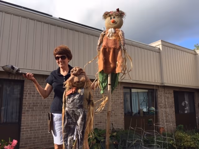 A woman wearing sunglasses and a black shirt is standing outside in front of a brick building, holding a garden tool. She is next to two scarecrows, one taller and one shorter, both dressed in rustic clothing. There are plants and garden structures around them under a partly cloudy sky.