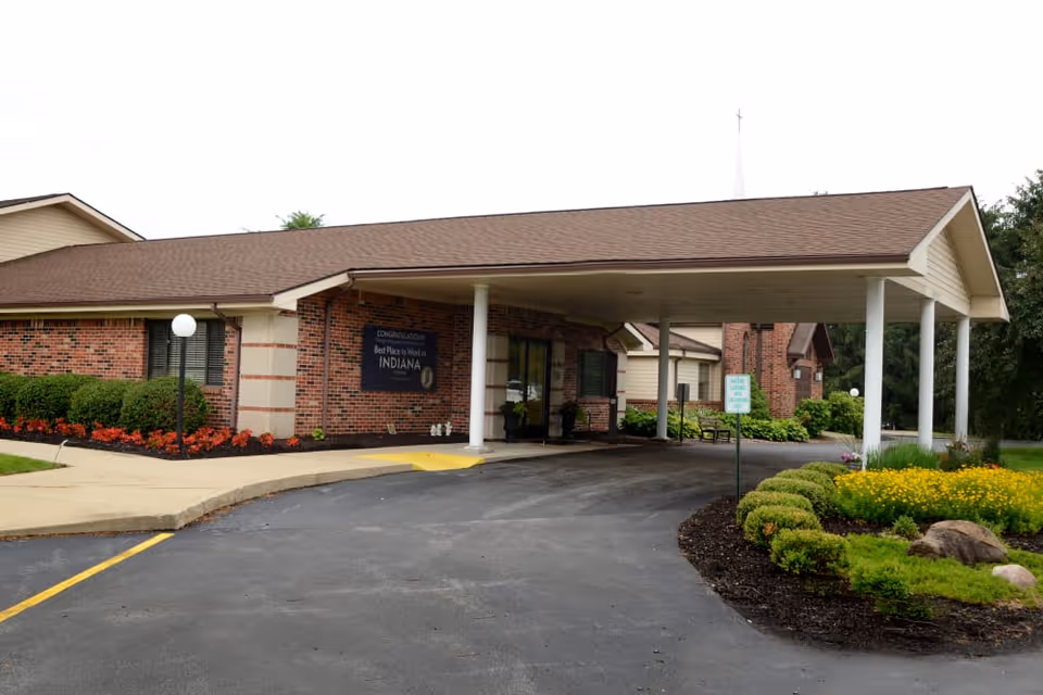 Front entrance of a brick senior living building with a covered driveway, columns, and landscaped flowerbeds.