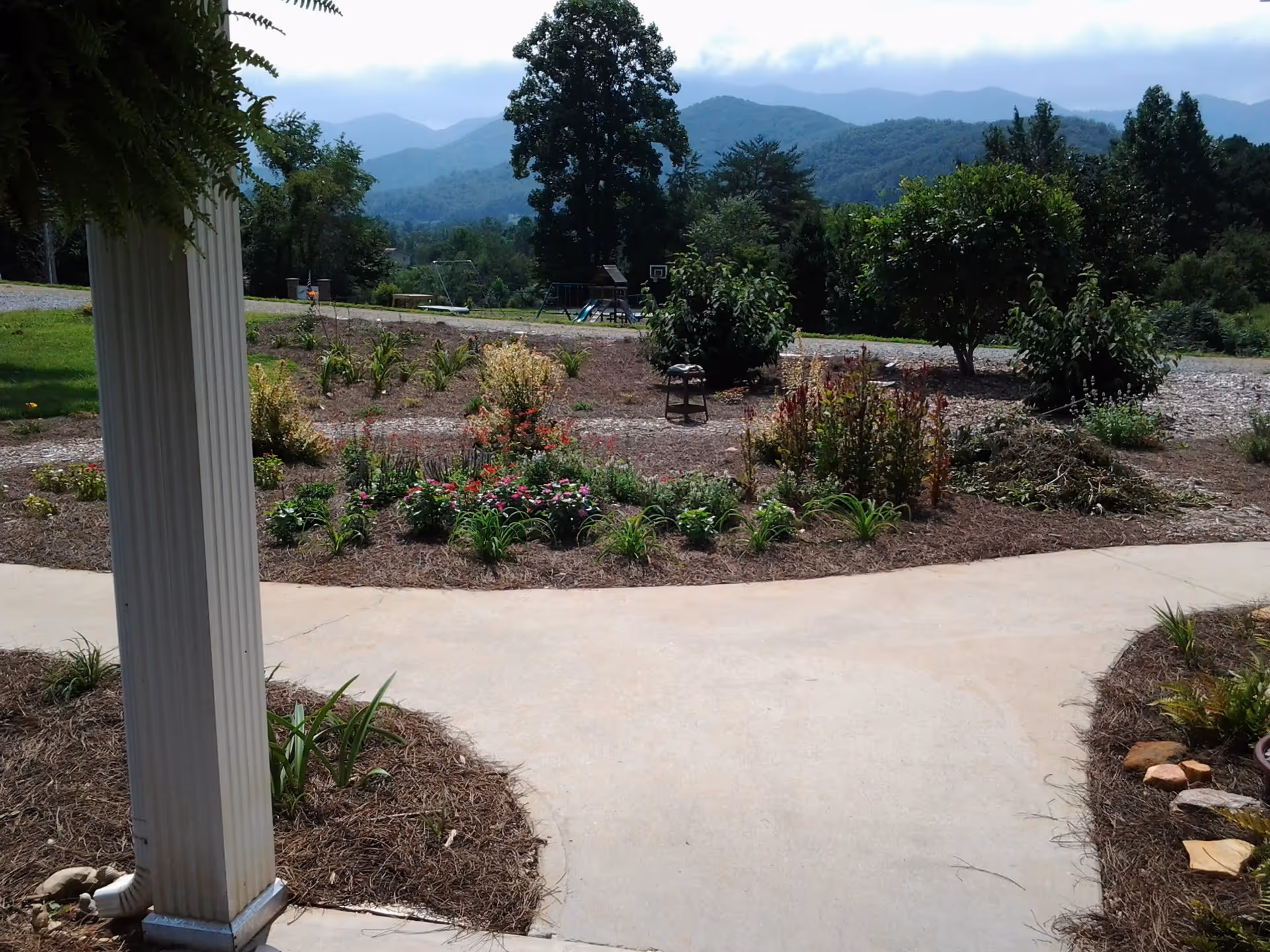 View of a landscaped garden area with various plants and flowers, a concrete pathway, and a large tree in the background. Beyond the garden, there is a playground with a swing set and slide, and mountains visible in the distance under a partly cloudy sky.