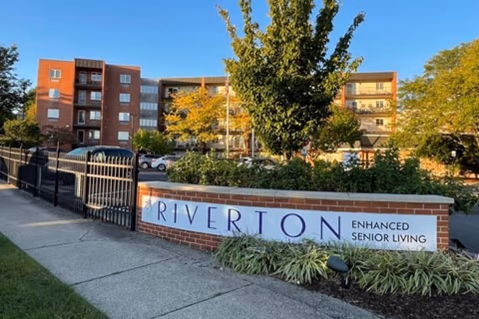 Exterior view of Riverton Enhanced Senior Living facility showing a brick sign with the facility name, a sidewalk, a black metal fence, trees, and a multi-story building in the background under a clear blue sky.
