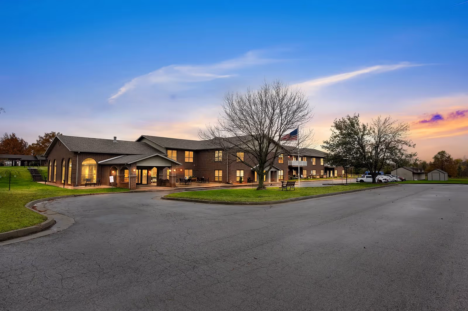 Exterior view of a two-story brick building at dusk with a curved driveway, a few parked cars, leafless and leafy trees, and an American flag on a flagpole in front of the building.