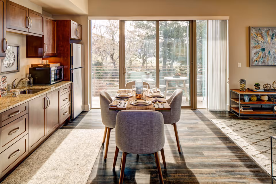 A bright dining area with a wooden table set for four people, surrounded by upholstered chairs. To the left is a kitchen with wooden cabinets, a granite countertop, a sink, a microwave, and a refrigerator. Large sliding glass doors in the background open to a balcony with outdoor seating and trees visible outside. To the right, part of a living room area with a rug, a console table, and a colorful abstract painting on the wall is visible.