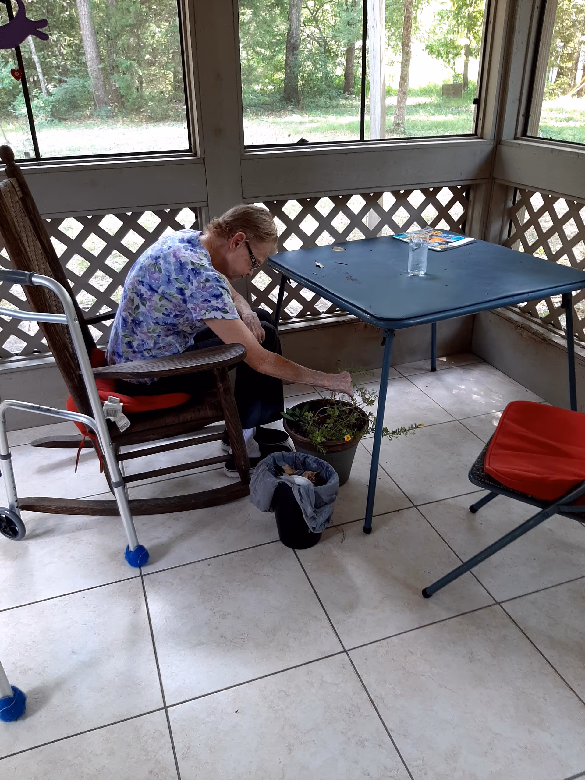 An elderly woman sits in a rocking chair on a screened porch tending a potted plant next to a folding table and a walker.