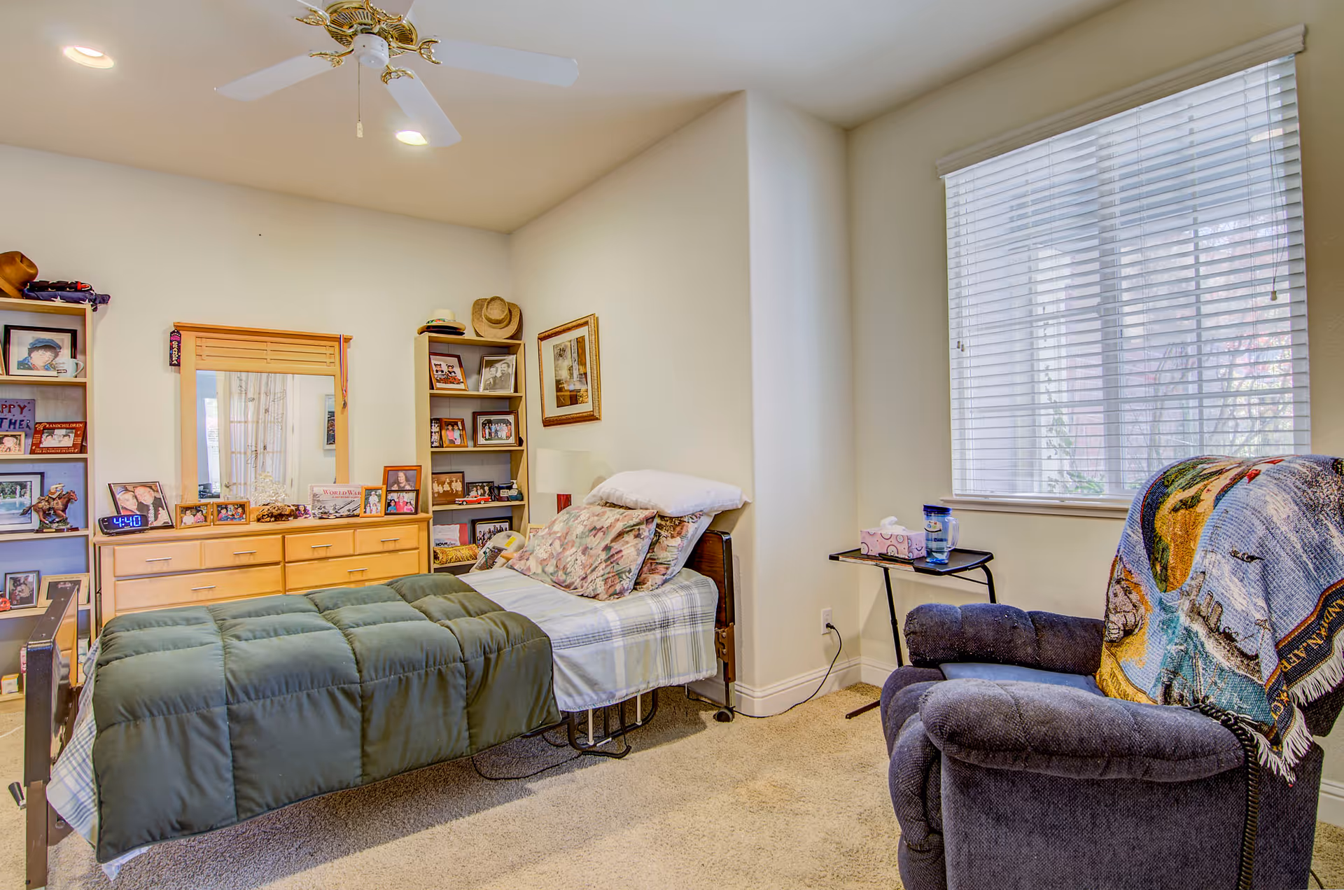 Cozy furnished bedroom with a single bed, dresser with photos, shelving, and a recliner by a window.