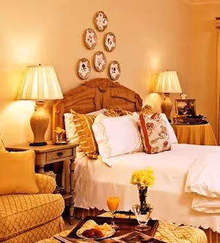 A cozy bedroom with a wooden headboard bed adorned with multiple pillows and a white bedspread. Two bedside tables each hold a lamp with a beige shade. Above the bed, decorative plates are arranged on the wall. In the foreground, there is a cushioned armchair and a small table with a vase of yellow flowers and a tray with food and drink.