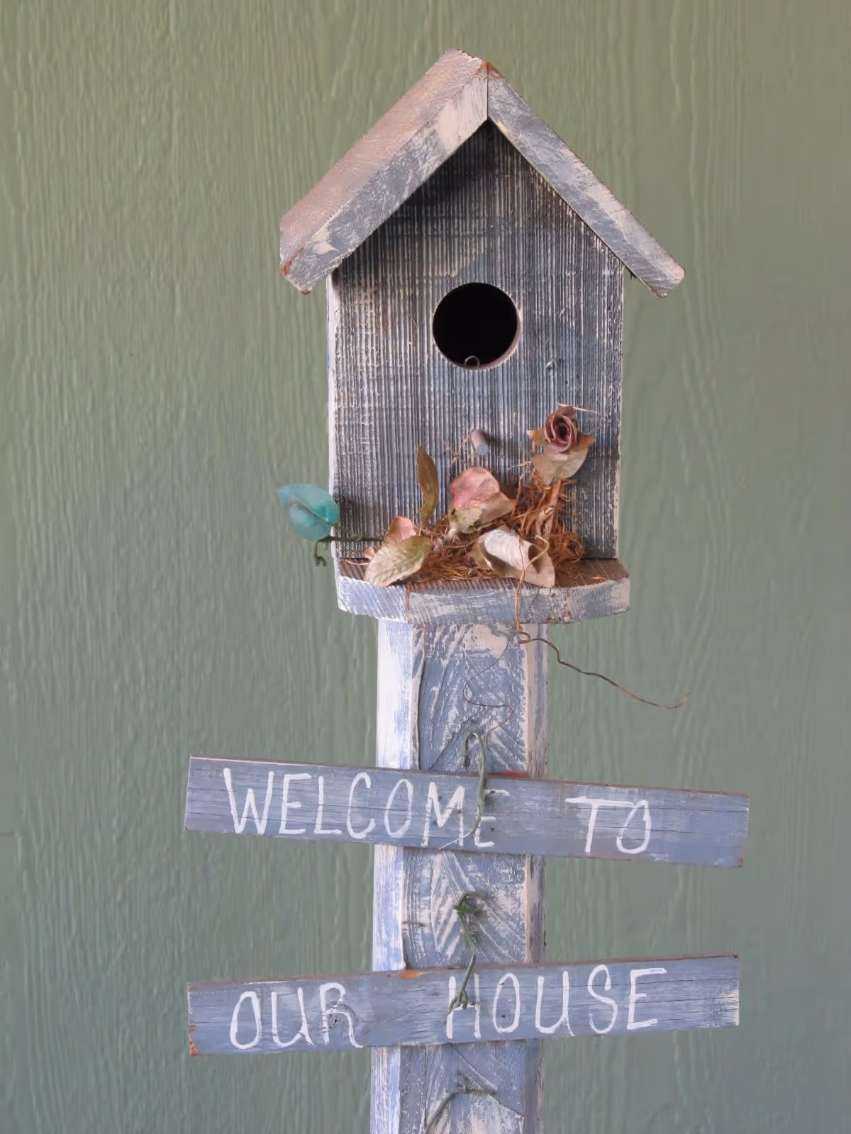 A rustic wooden birdhouse on a post decorated with dried flowers and signs reading 'Welcome to our house' against a green wall.