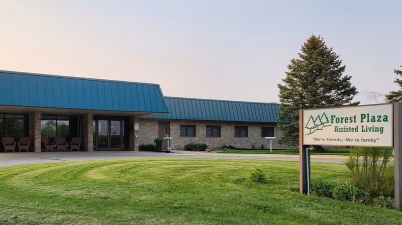 Exterior view of Forest Plaza Assisted Living facility with a green lawn in front, a building with a blue roof, and a sign that reads 'Forest Plaza Assisted Living - We're friends--We're family'.