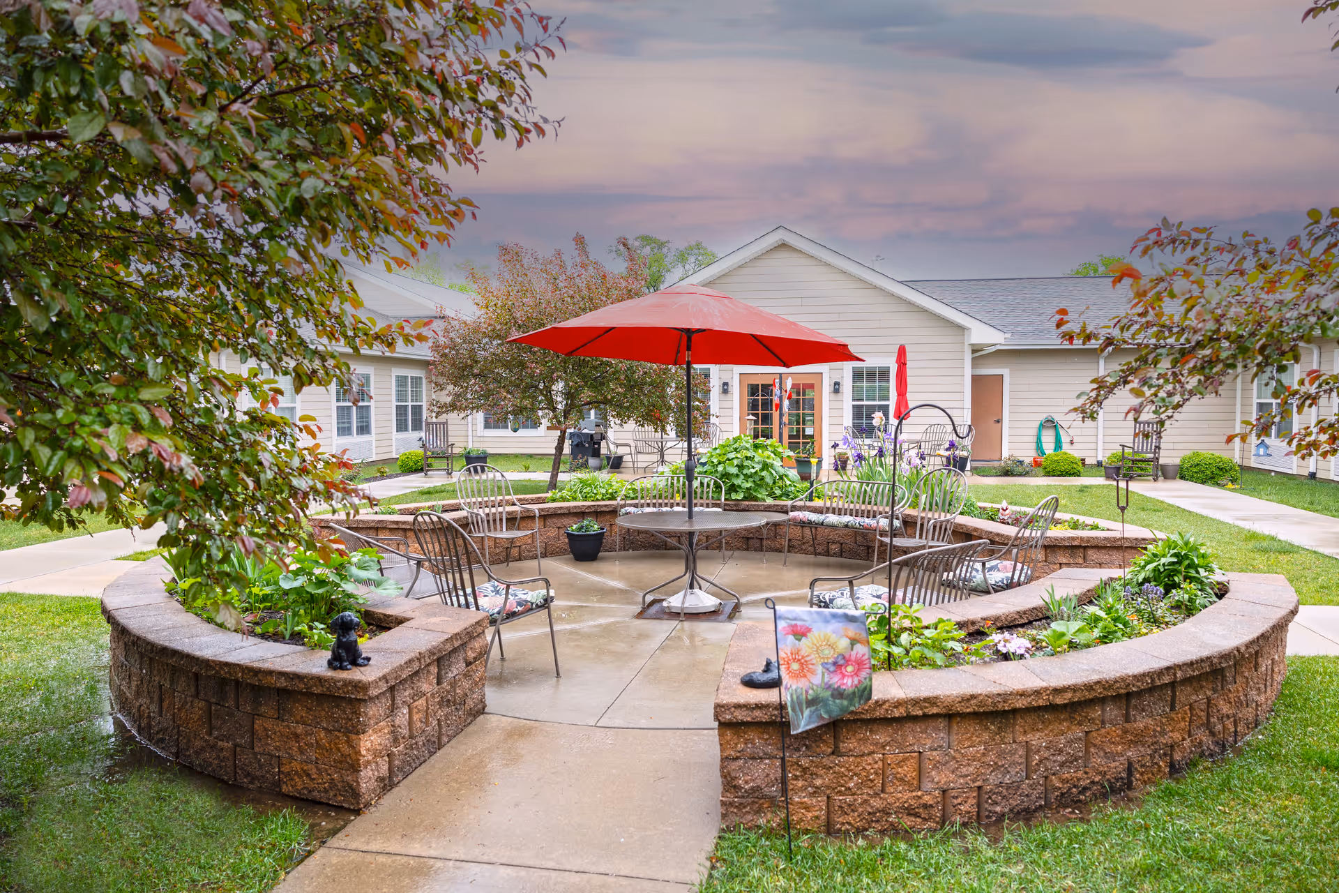 Outdoor courtyard area with a circular stone seating wall surrounding a patio table with a red umbrella. Several metal chairs with floral cushions are arranged around the table. The courtyard is landscaped with green plants and flowers, and there are trees and a beige building in the background under a cloudy sky.