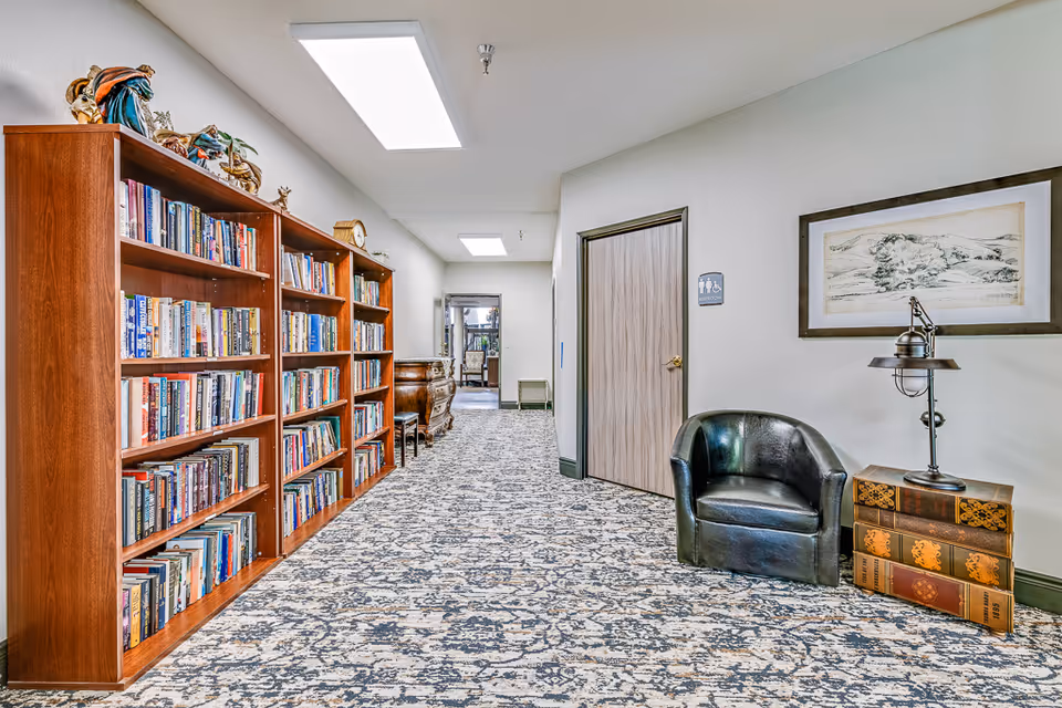 A hallway in a senior living facility with carpeted floor and white walls. On the left side, there are wooden bookshelves filled with books and decorative items on top. On the right side, there is a black leather chair next to a small table designed to look like stacked vintage books with a lamp on it. A framed landscape artwork hangs above the chair. At the end of the hallway, there is a wooden door with a restroom sign indicating accessibility. Further down the hallway, there is a glimpse of another room with furniture.