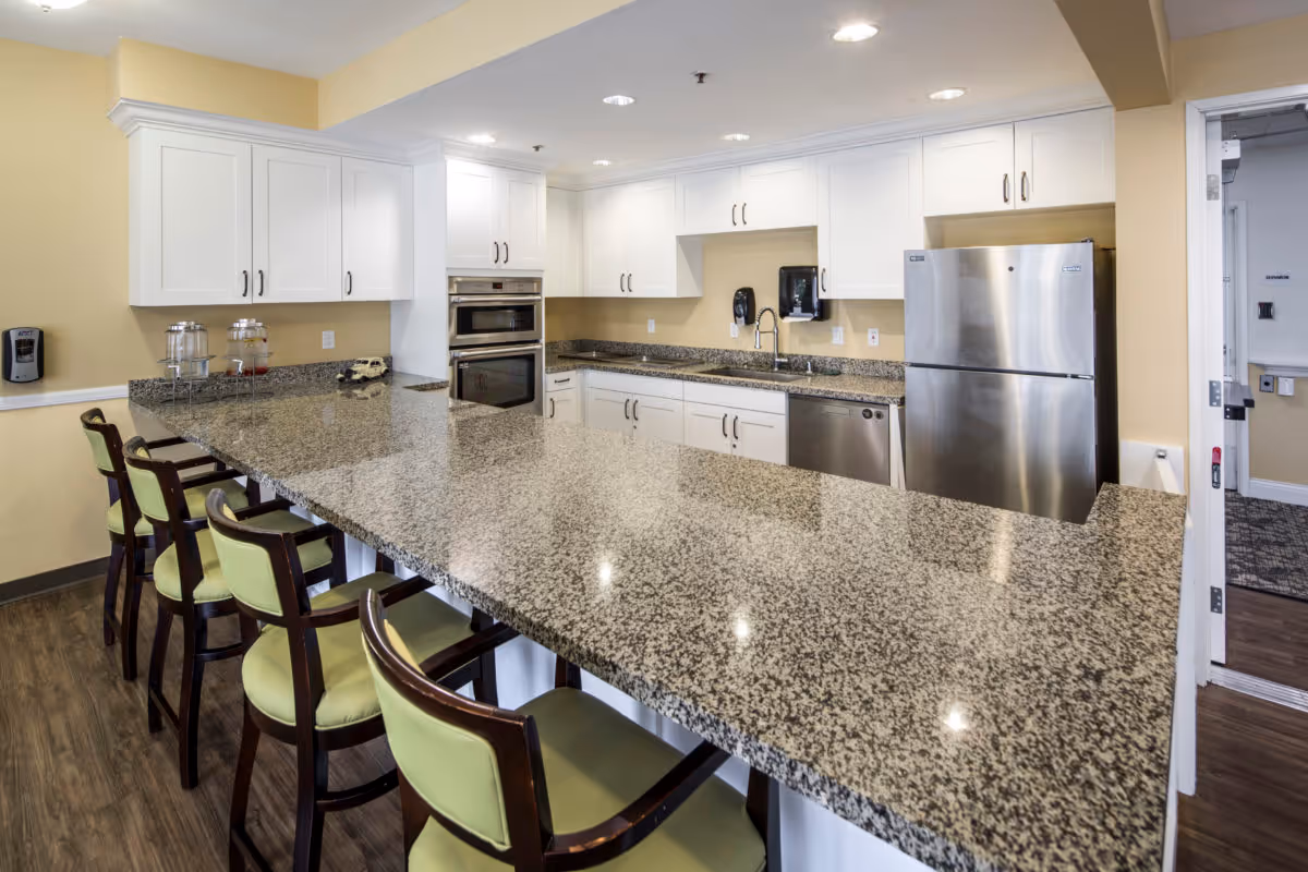 A modern kitchen with a long granite countertop island surrounded by six wooden chairs with green cushions. The kitchen features white cabinets, a stainless steel refrigerator, built-in oven, dishwasher, and a sink with a faucet. The walls are painted light yellow and the floor has a wood finish.