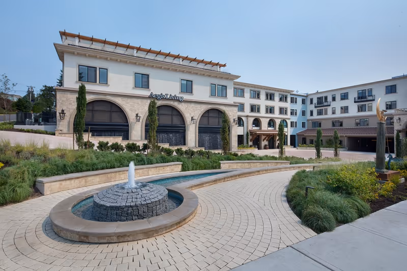 Front exterior of Aegis Living Kirkland Waterfront showing a multi-story building with arched entrances, a curved fountain, and landscaped courtyard.