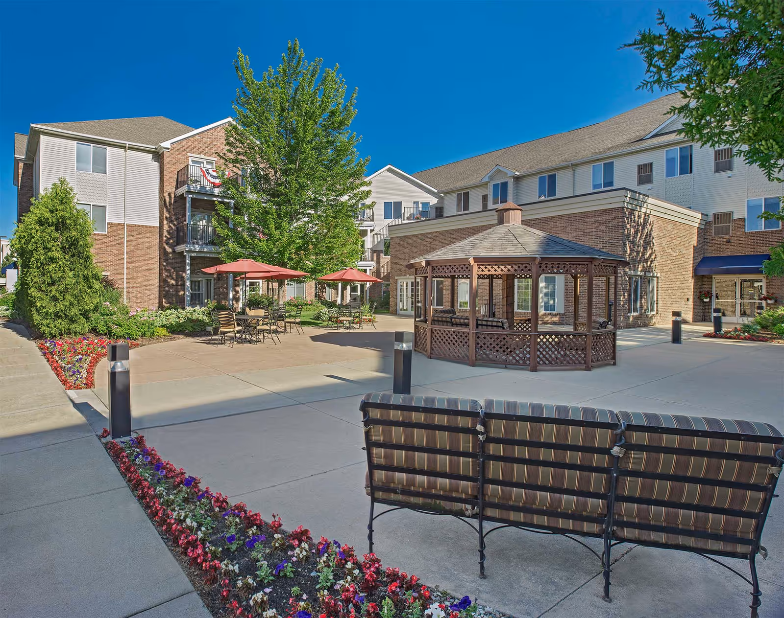 Outdoor courtyard area of a senior living facility with a wooden gazebo, patio tables with red umbrellas, benches, flower beds, and a multi-story building in the background under a clear blue sky.