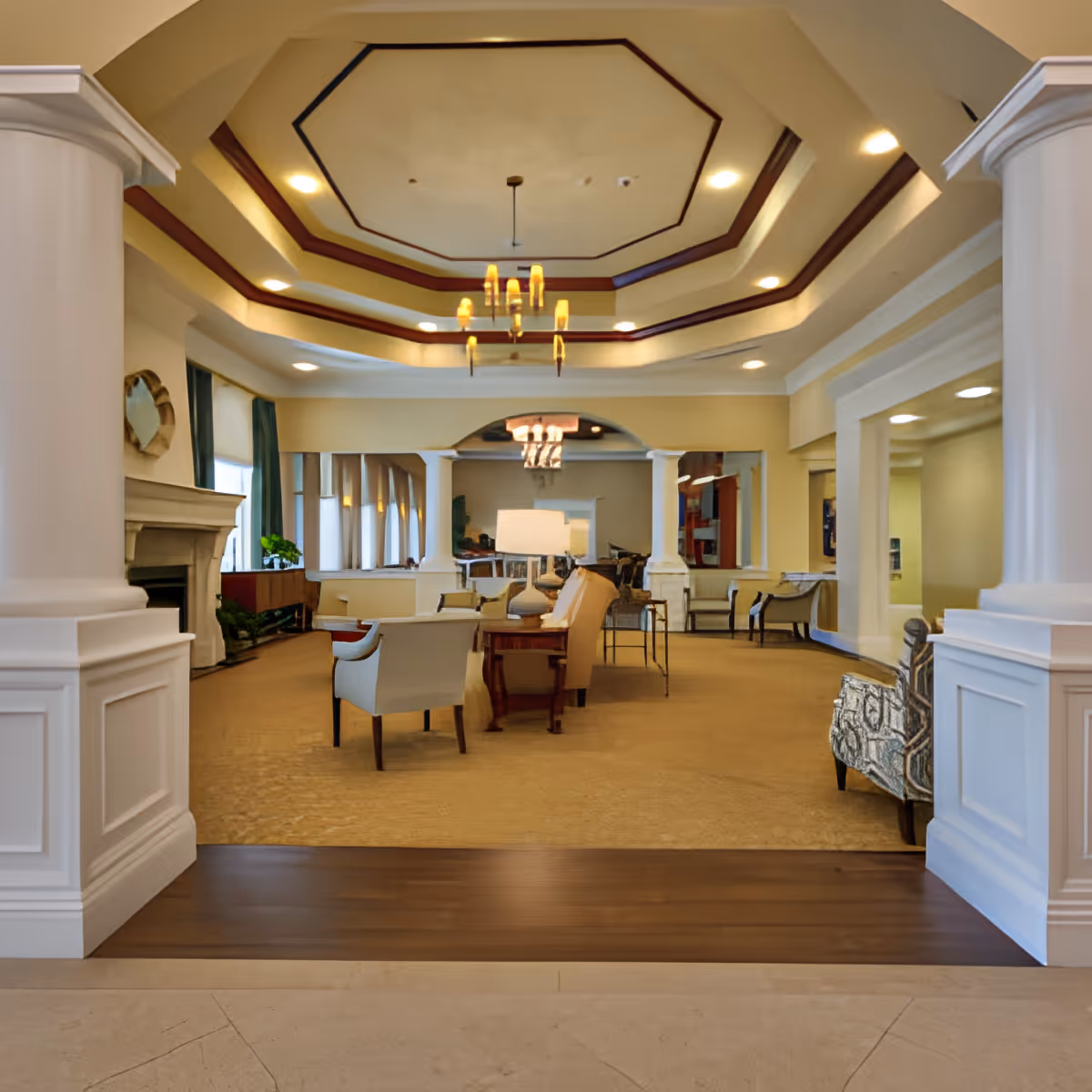 Open, well-lit senior living common room with couches, chairs, columns, and a decorative recessed ceiling with chandeliers.