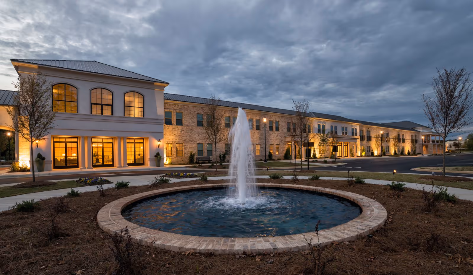 Exterior view of The Blake at Flowood senior living facility at dusk, featuring a large two-story building with illuminated windows and a central water fountain in the foreground surrounded by landscaping and a paved driveway.