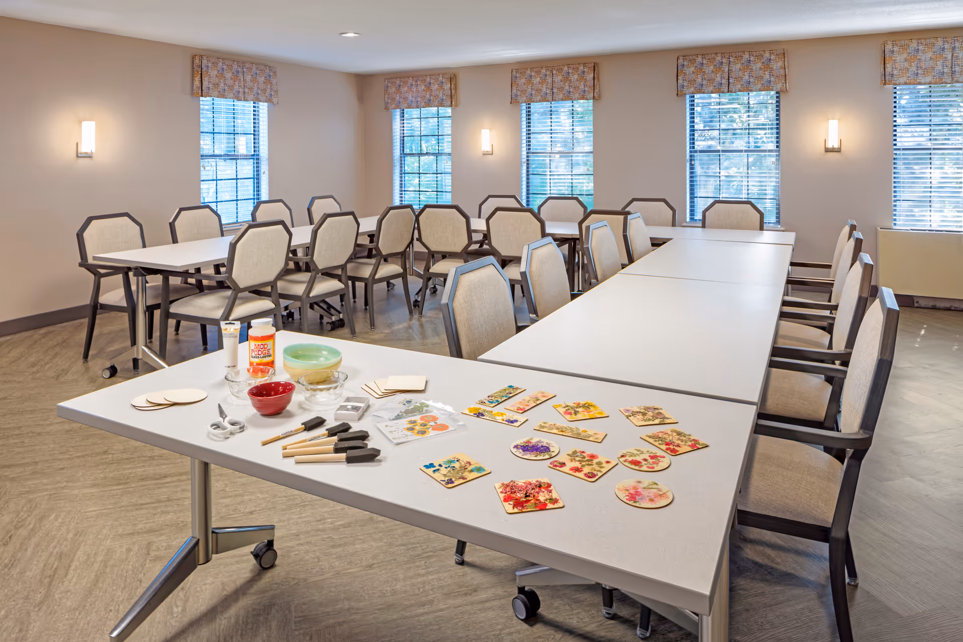 A well-lit activity room with several tables and chairs arranged in a U-shape. On the closest table, there are various craft supplies including scissors, paintbrushes, bowls, Mod Podge glue, and decorated wooden pieces with floral designs. The room has multiple windows with blinds and patterned valances, and wall-mounted lights.