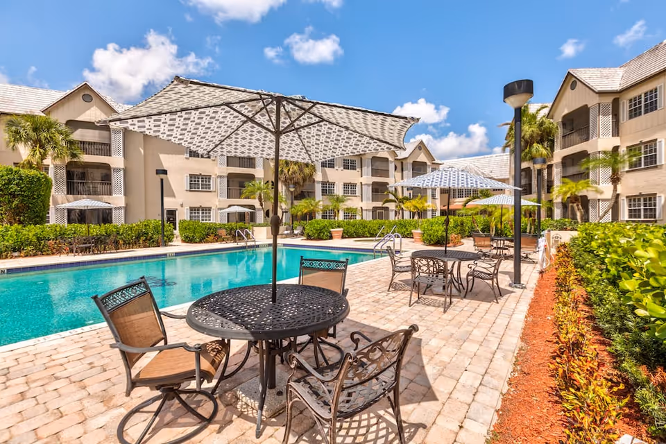 Outdoor pool and patio area with umbrella-shaded tables and chairs in front of a multi-story apartment-style building.