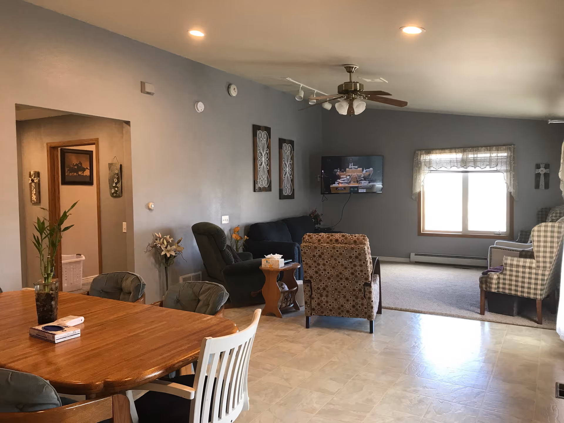 A cozy living room and dining area in an assisted living facility. The dining area features a wooden table with chairs, a plant, and some books. The living room has a variety of armchairs and a sofa arranged around a wall-mounted TV. The walls are painted gray, and there is a window with a sheer curtain letting in natural light. Decorative wall art and a ceiling fan with lights are also visible.