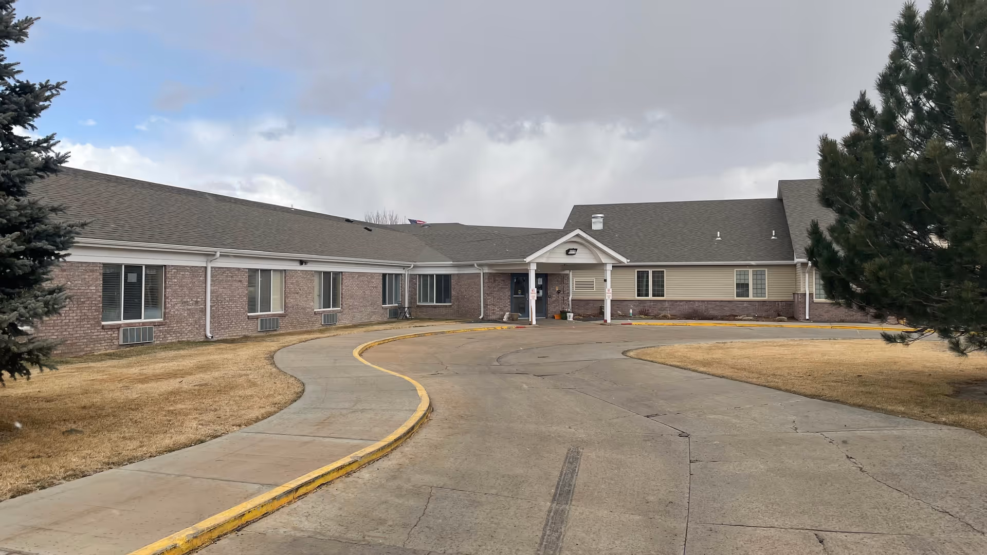 Front entrance of a single-story brick-and-siding senior living building with a curved driveway and grassy lawn under a cloudy sky.