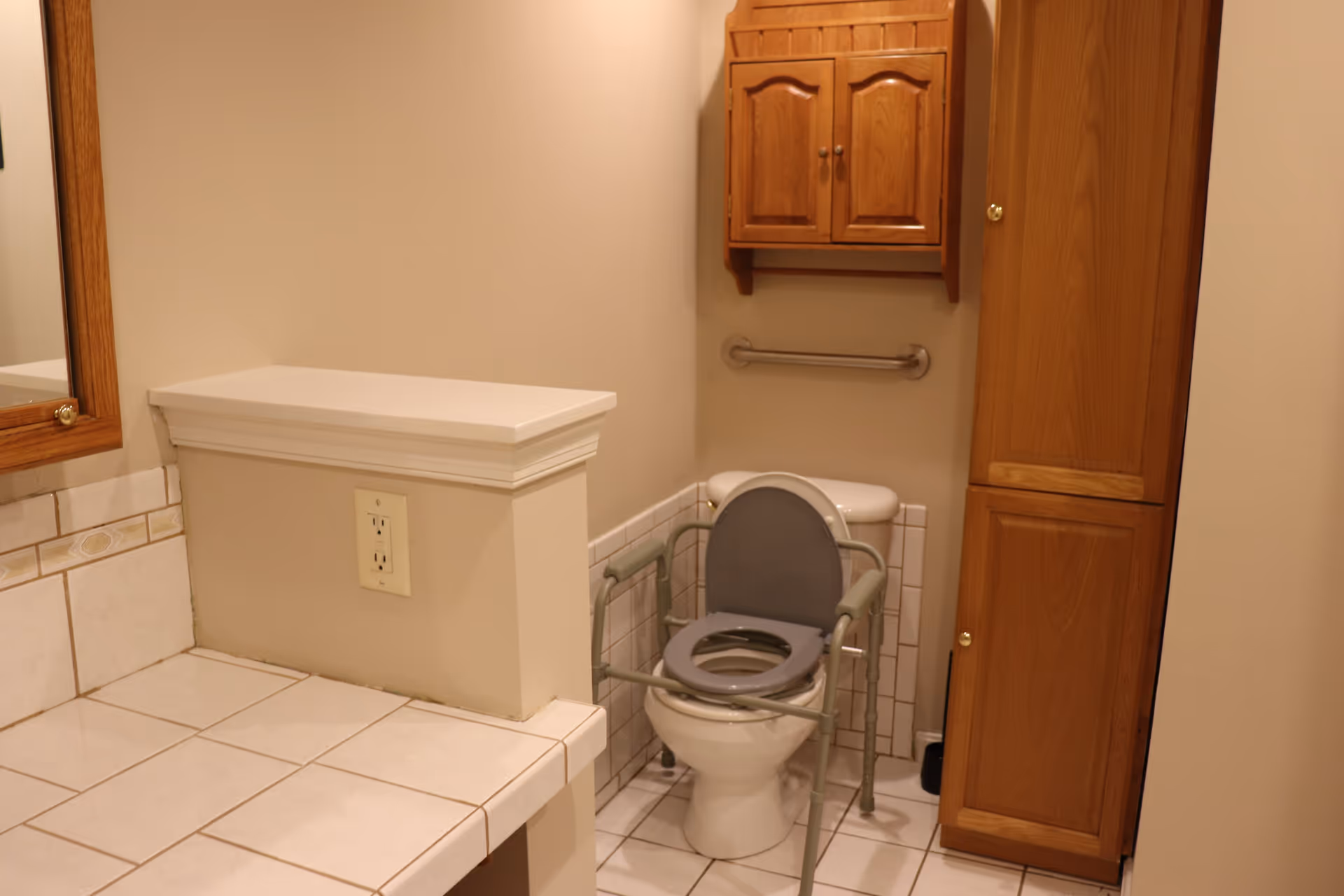 A bathroom with a white toilet equipped with a gray raised toilet seat and safety rails. Above the toilet is a wooden cabinet mounted on the wall and a metal grab bar. To the right of the toilet is a tall wooden storage cabinet. The floor and countertop are tiled in white, and there is an electrical outlet on the wall near the countertop.