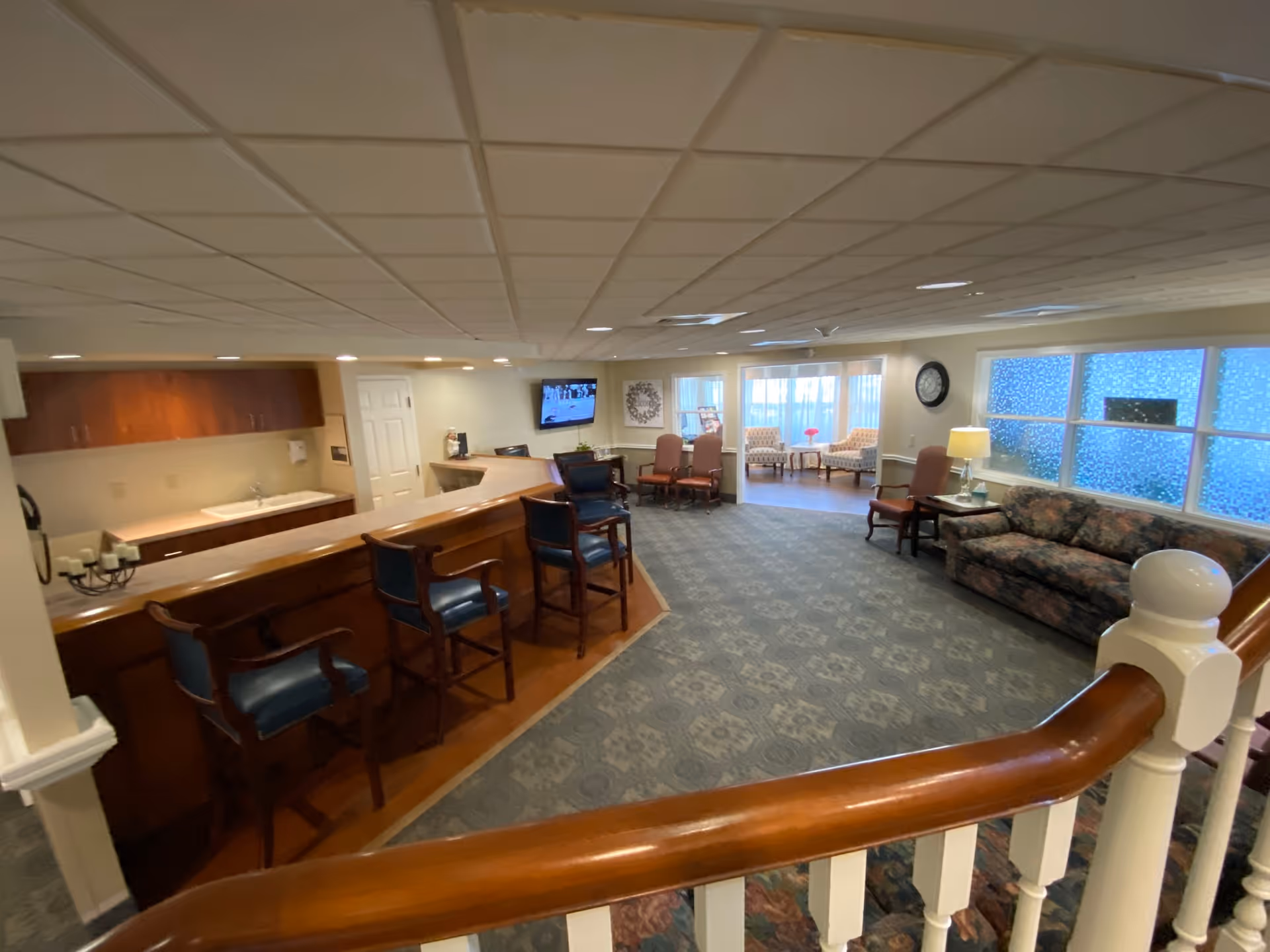 Interior view of a senior living facility common area with a wooden counter and several high chairs on the left, a carpeted floor, a floral-patterned couch on the right, and additional seating with armchairs near large windows in the background. A TV is mounted on the far wall, and the area is well-lit with ceiling lights.